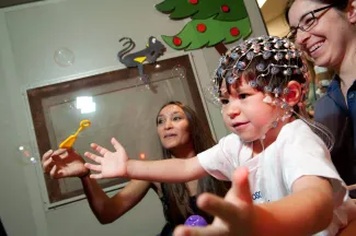 A baby wearing an EGG machine on his head reaches for bubbles that a woman is blowing.