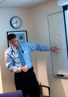 Male instructor wearing shirt and tie points to diagram on white board