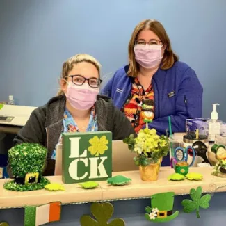 two women wearing masks behind a desk of st. patricks day decor