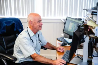 an older white male doctor sitting in front of a desk with a couple monitors