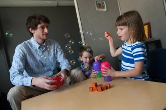 A girl blows bubbles at a table while a man sits and talks with her while another child plays between them.