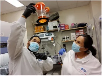 man and woman in white lab coats and blue face masks in a lab looking into a orange and clear container