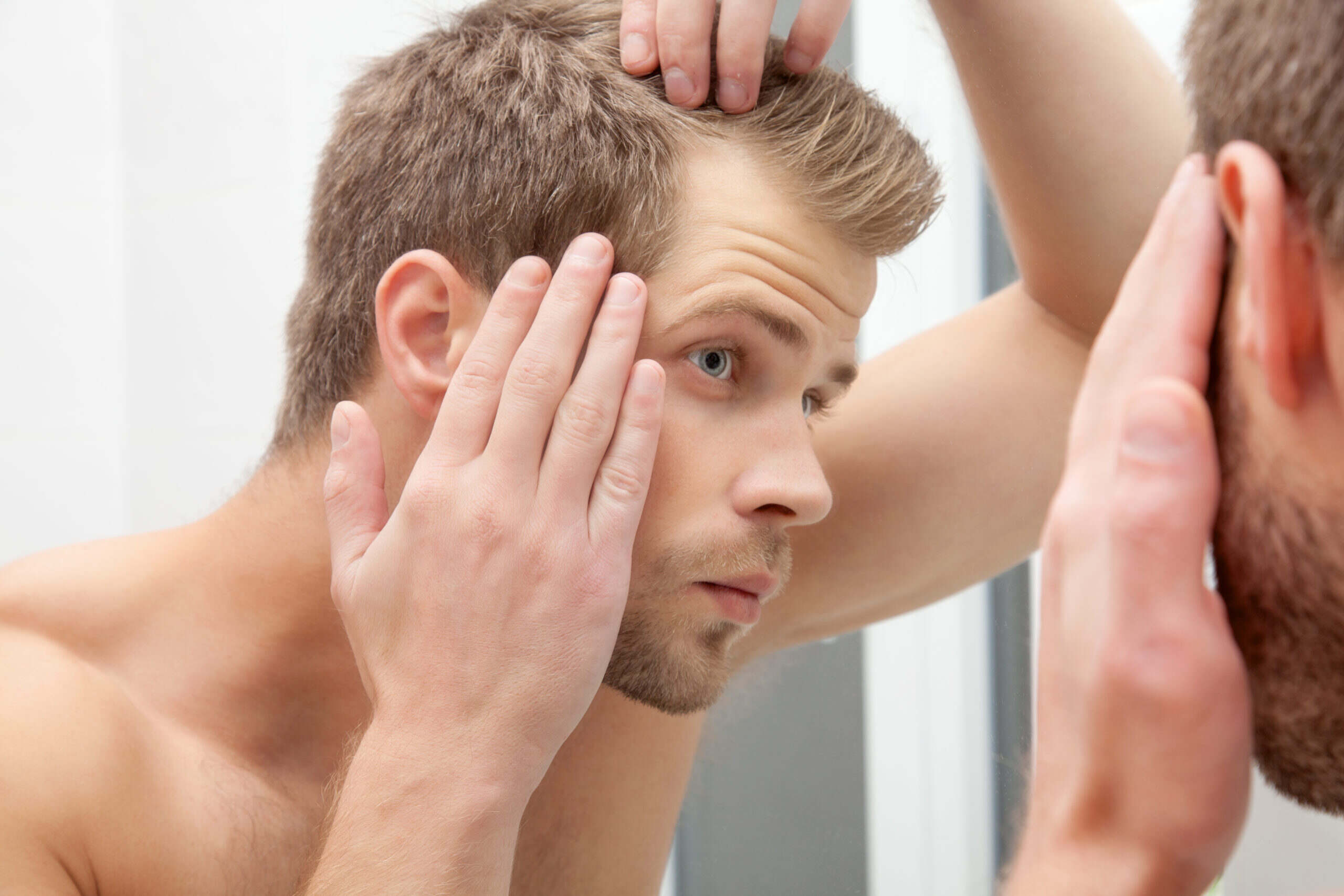 Young man assessing his hair loss in the mirror and thinking about hair restoration treatments.