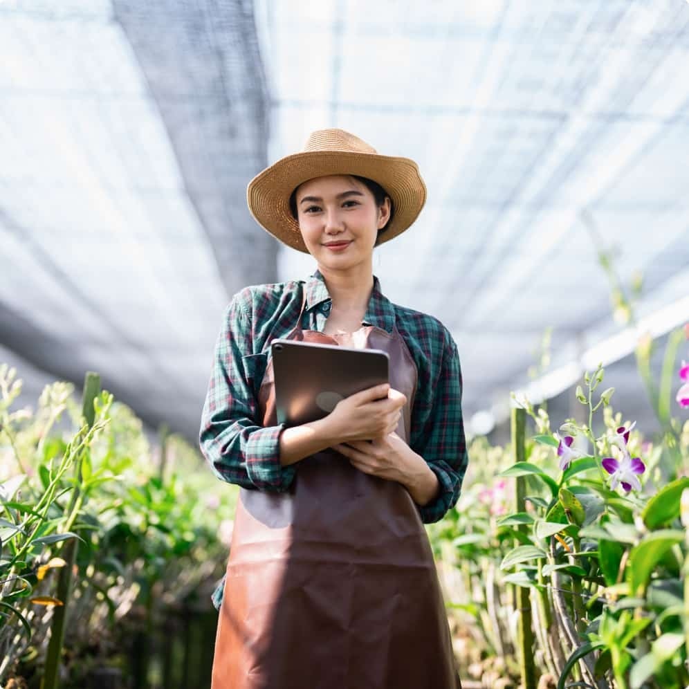 A woman softly smiles at the camera, wearing a sun hat while holding a tablet inside of a greenhouse. 