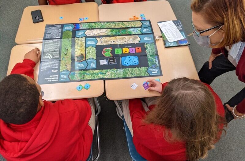 To help raise geography awareness, Mary Shorse plays a board game with students. The game illustrates with Landsat imagery six unique river systems across the United States and allows students to see how geography influences where they get their water from and how water is used, both for drinking and for making certain products. 