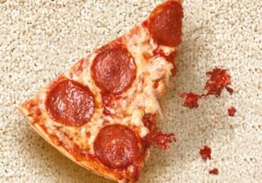 Pizza slice sitting on carpet with sauce stains. 