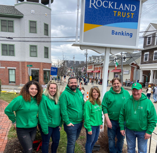 A group of six Rockland Trust employees smiling together wearing matching green sweatshirts as they volunteer at Boston's annual Saint Patrick's Day parade.