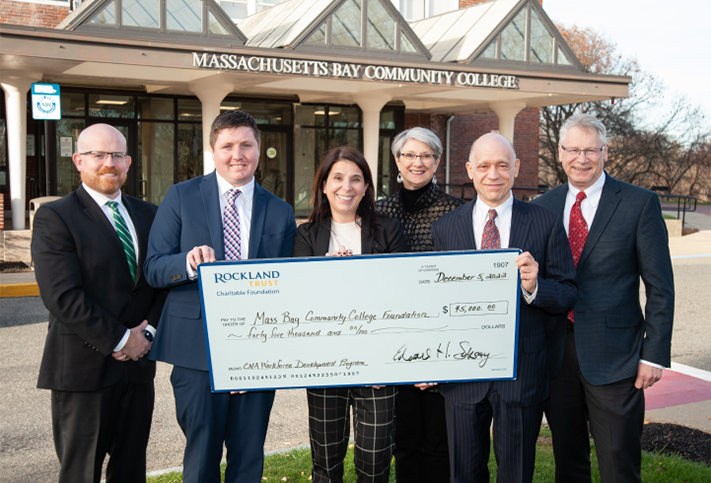 Members of Rockland Trust's Charitable Giving team smiling together, holding a large check for Massachusetts Bay Community College.