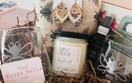 Holiday gift box containing a wine glass, apple cider scented candle, aromatic spray, and handmade earrings, all sold by The White Belle.