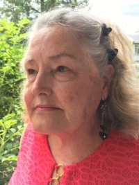 Image 1. An older white woman looking towards the left of the camera. She is wearing a pink shirt and has grey hair that is pinned back. She is outdoors in front of green bushes.