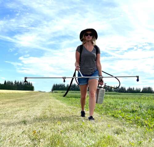 A woman wearing a large brimmed hat walks along a field with a 4-nozzle sprayer and a canister