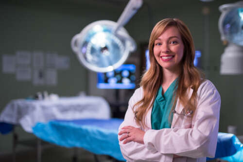 A woman with long blond hair stands in scrubs in front of an operating table smiling at the camera.