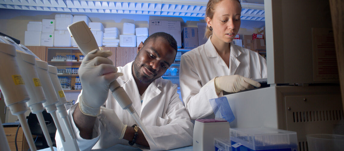 Male student takes sample in lab, while female student works at computer; both in lab coats