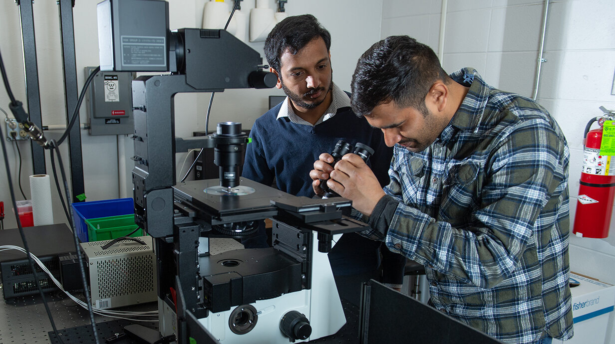 A man looks through a microscope in a laboratory while another man observes.