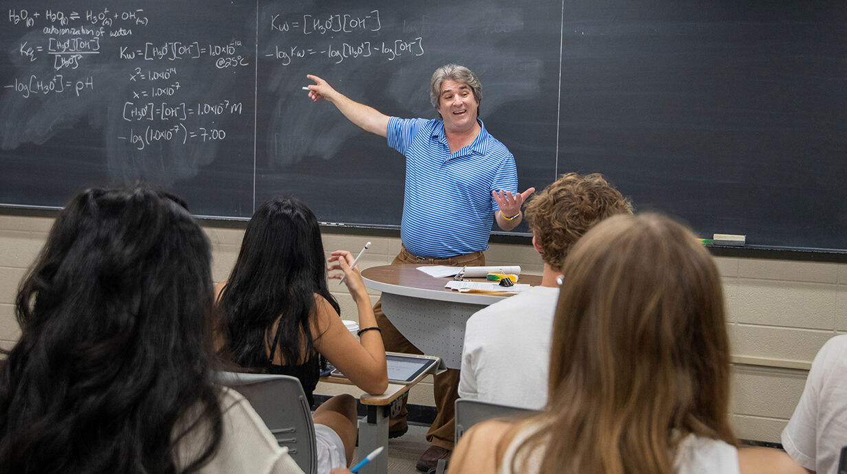 A man points to equations on a chalkboard at the front of a classroom.