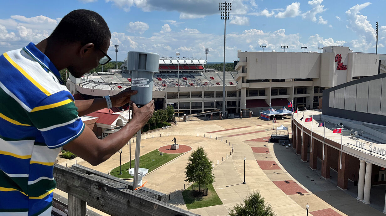 A researcher adjusts an air senors on a rooftop overlooking a football stadium.