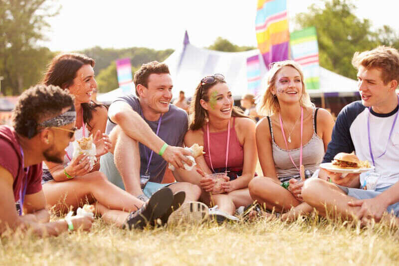A group of friends sit in the grass eating food at a festival.