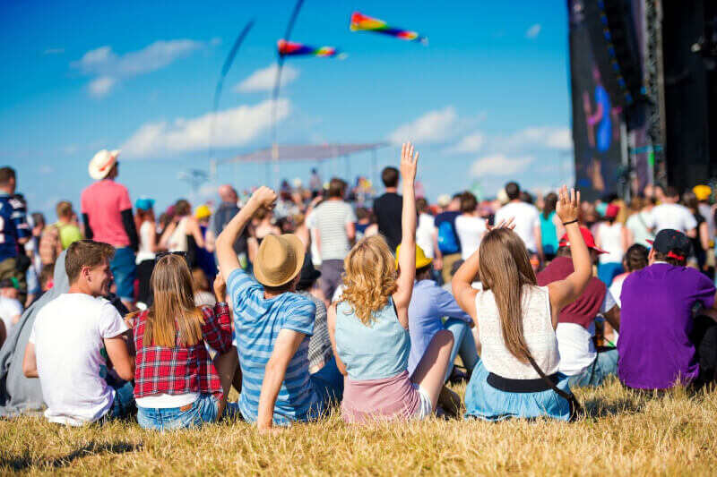A line of people sit in the grass at a music festival behind a crowd of people.
