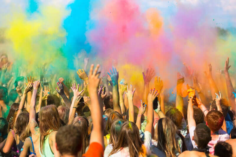 A crowd of people at a festival with their hands raised as clouds of colorful smoke billow over them. Some of the raised hands are colorful, as well.