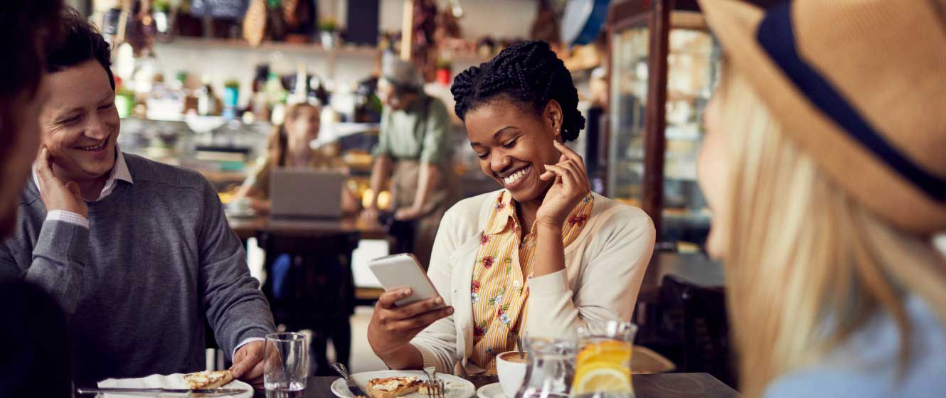 Woman sitting at table in restaurant with friends while smiling and looking at her smart phone