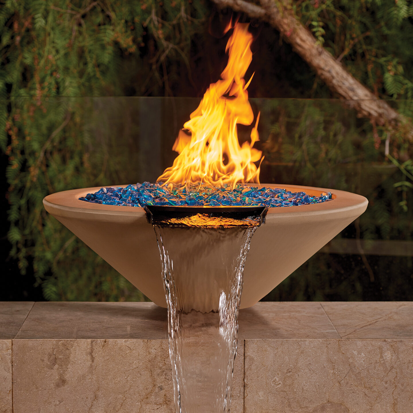 A fire bowl with colorful glass pieces and flames, featuring a water spillway, on a stone pedestal.