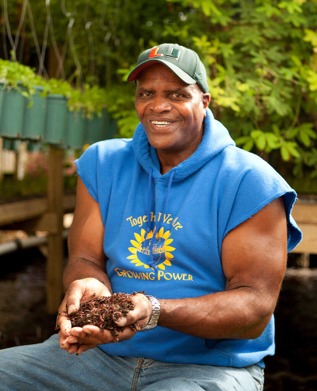 Photo of Will Allen wearing a blue sweatshirt with the sleeves cut off that reads "Together we are growing power" and features a graphic of a sunflower. He is holding a handful of soil with a garden in the background.