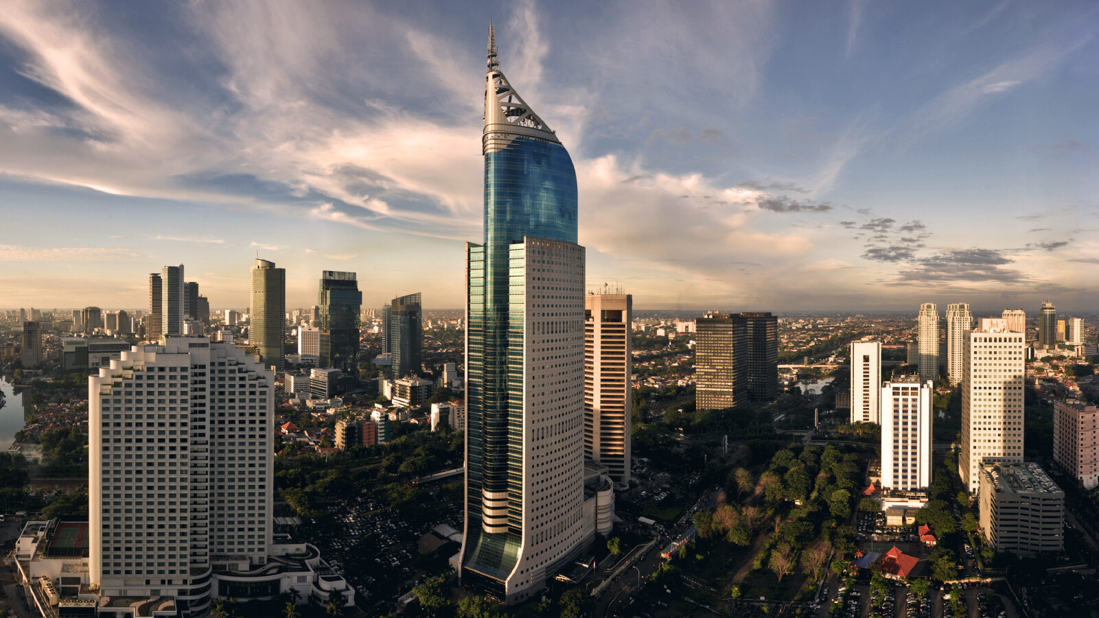 Jakarta city skyline at sunset featuring the iconic BCA Tower surrounded by modern high-rise buildings under a partly cloudy sky.