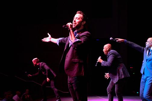 A member of THE COMPANY MEN singing on stage while three other members sing behind him. 