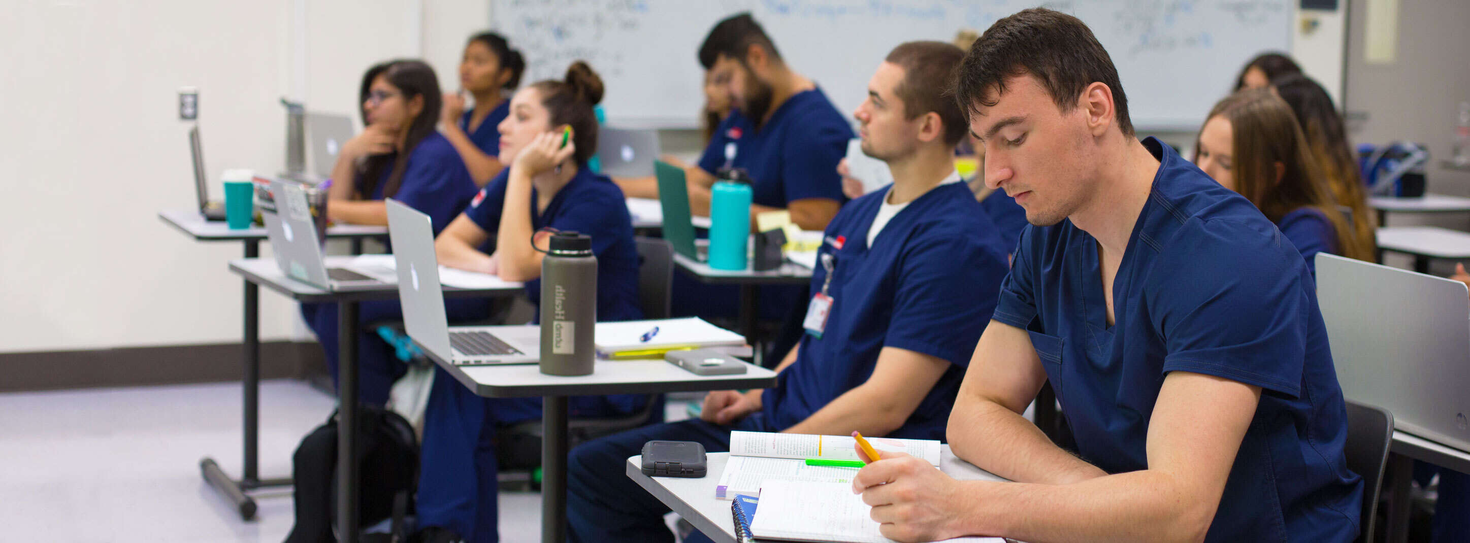 A group of students wearing scrubs and sitting at desks while taking lecture notes.