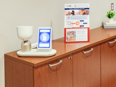 ElliQ is designed to foster independence and improves well-being and social connectedness Five people stand together in an office setting near a cabinet and coffee table. A wall-mounted display screen and seating area are visible in the background.